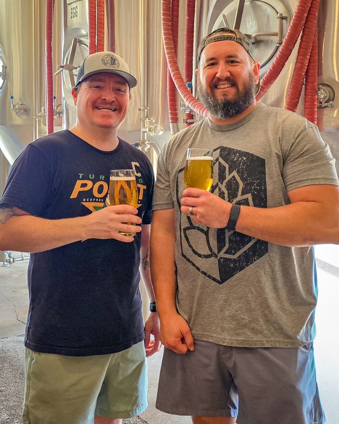 Two brewers in branded tees raising glasses of pale beer by the stainless fermenters at Turning Point Beer in Bedford, TX